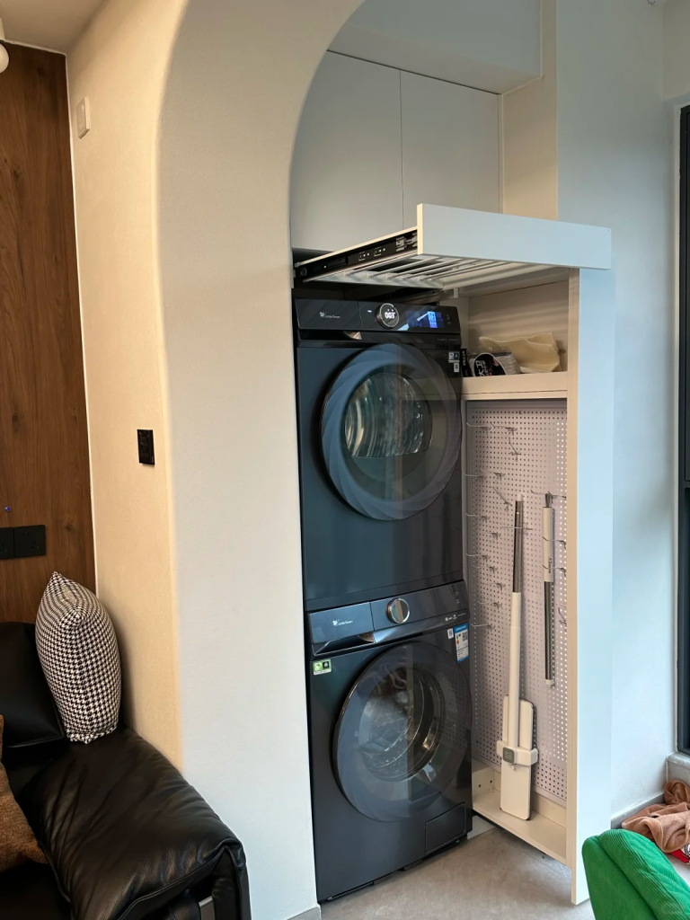 Stacked black washer and dryer in a narrow apartment alcove with a tall storage niche for cleaning tools.