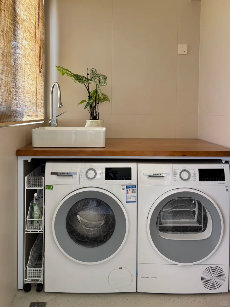 Side-by-side front-loading laundry machines with a slim pull-out wire storage rack beside a wooden counter and sink.