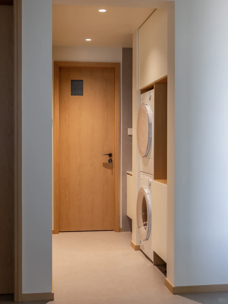 Stacked white washer and dryer fitted into a recessed hallway laundry area with pale built-in cabinetry.