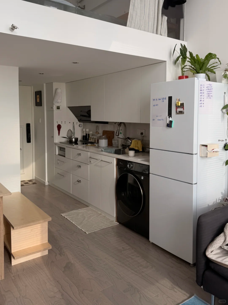 Black front-loading washing machine installed beneath a white kitchen counter in a compact open-plan apartment.