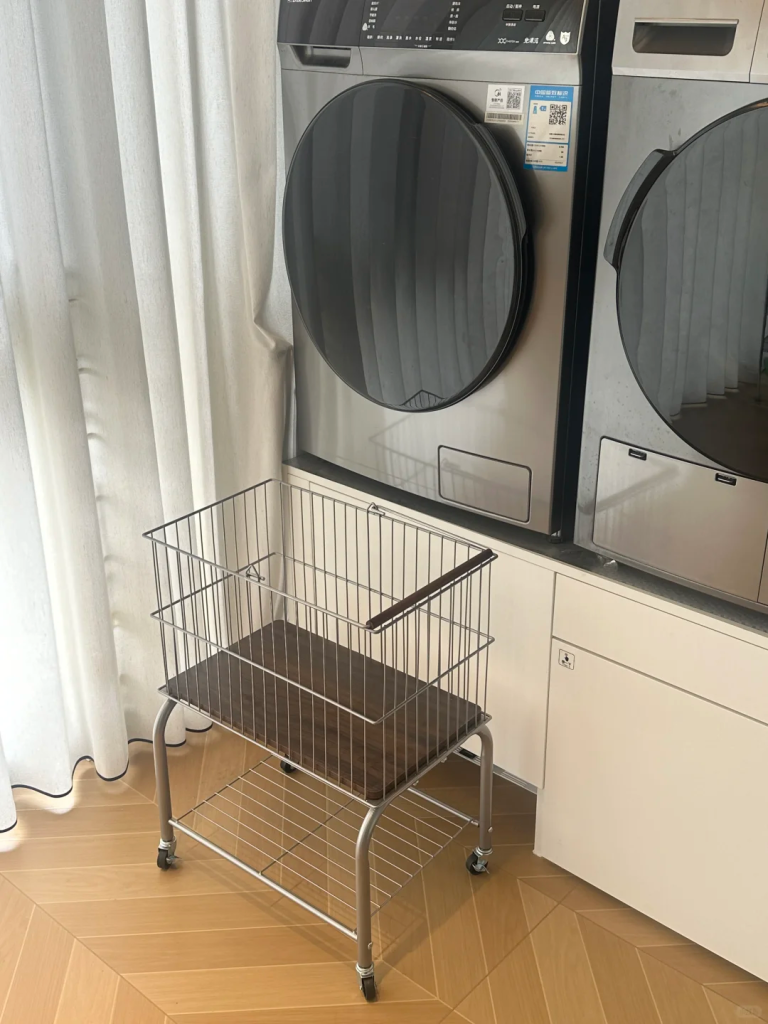 Metal rolling laundry basket with a lower storage shelf beside front-loading machines and white curtains.