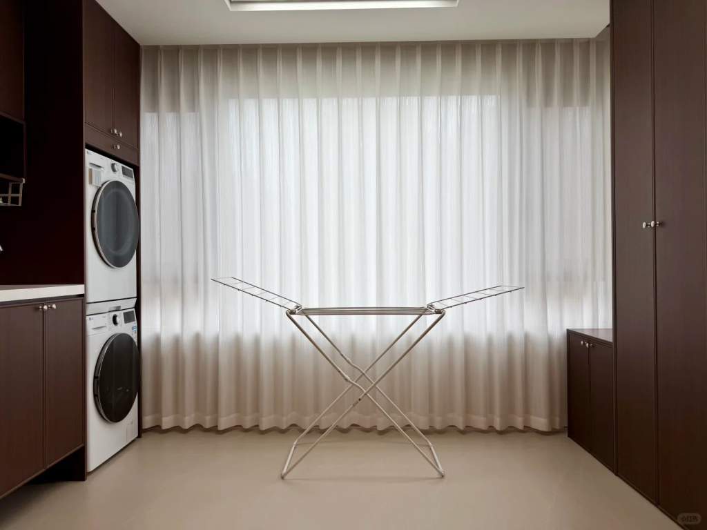 Freestanding winged clothes drying rack in a bright laundry room with stacked machines and large curtained windows.