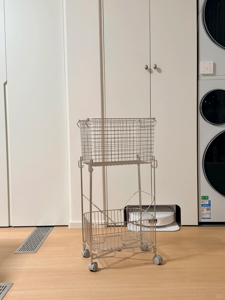 Two-tier metal rolling laundry basket standing in front of white cabinets beside stacked laundry machines.