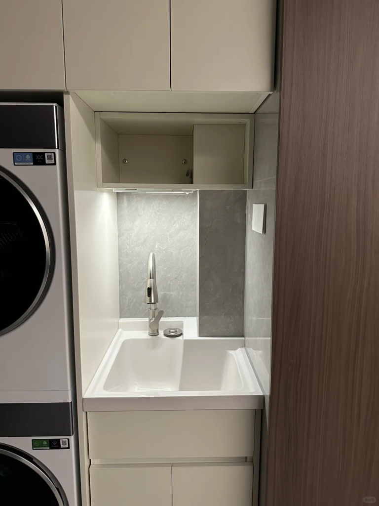 Compact white utility sink beside stacked laundry machines, illuminated by an under-cabinet task light.