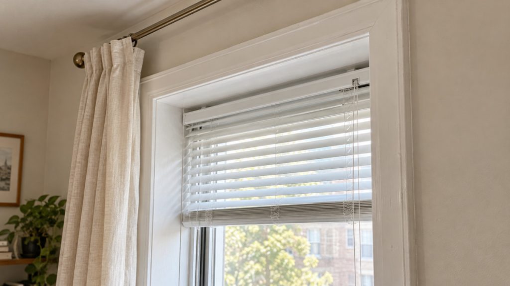 Close-up of a rental window showing the recessed horizontal blinds, white trim, and curtain placement.