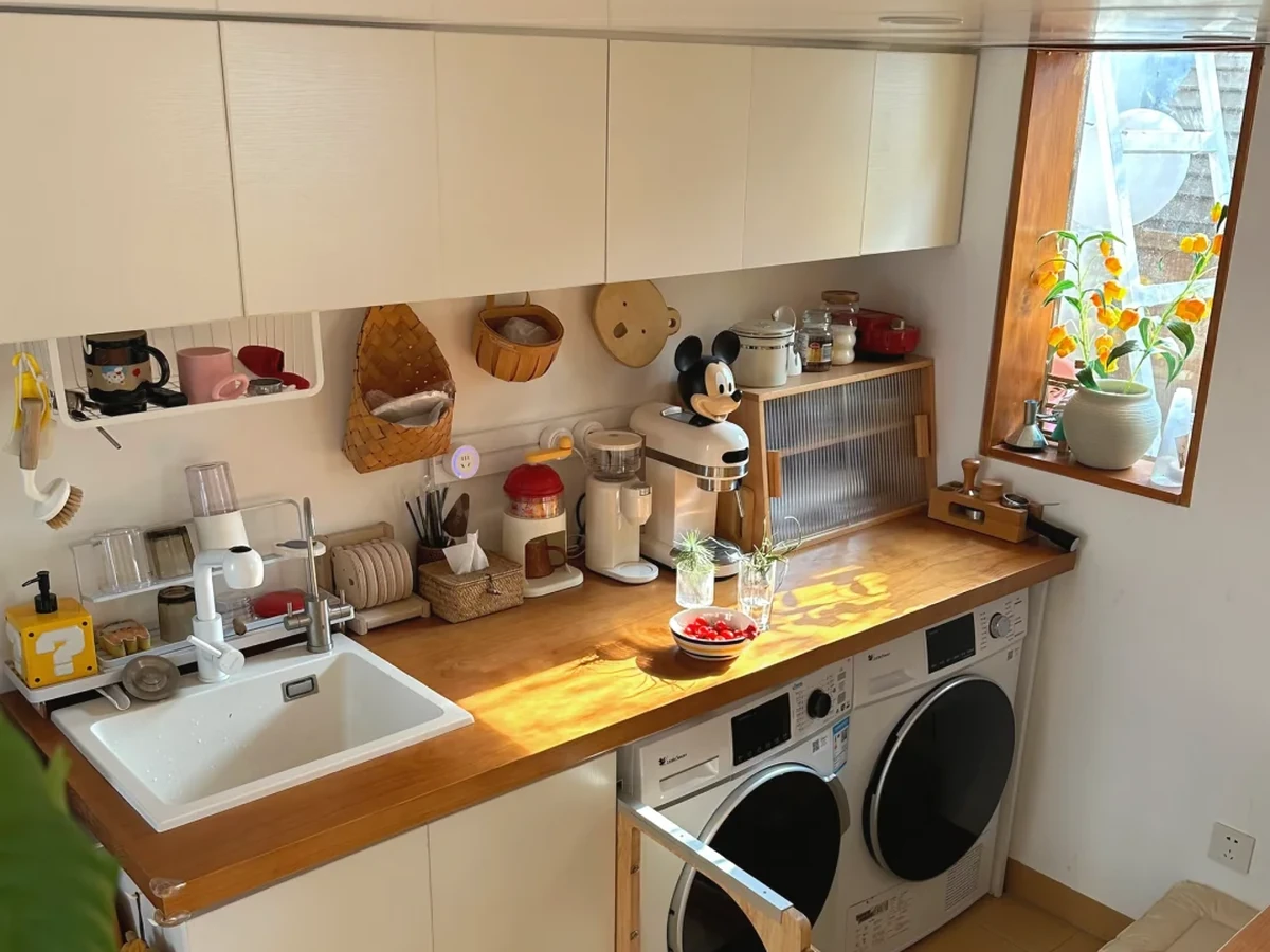 Small apartment laundry room with side-by-side washer and dryer, wooden countertop, overhead cabinets, and compact sink.